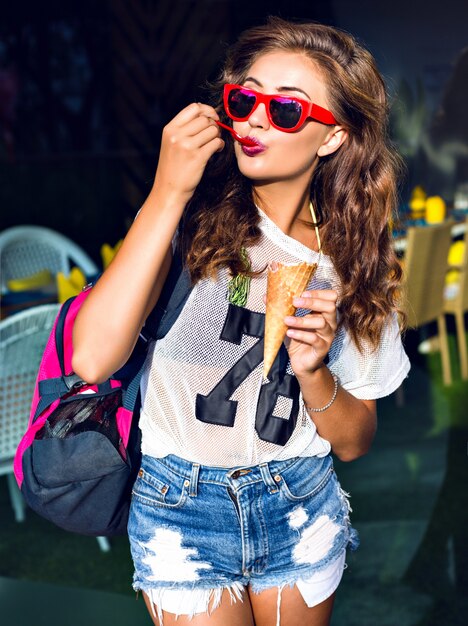 Beautiful young woman eating a big sweet ice cream in red sunglasses, shorts, sports bag on his shoulder, standing outside in summer.