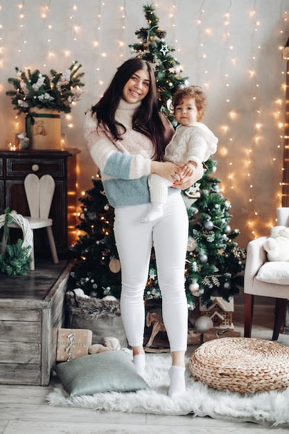 Beautiful young woman in fluffy sweater smiling while holding child and standing near Christmas tree