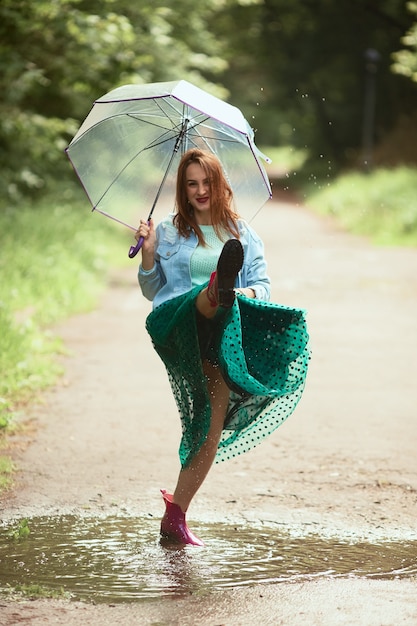 Beautiful young woman in green skirt has fun walking in gumboots on pools after the rain