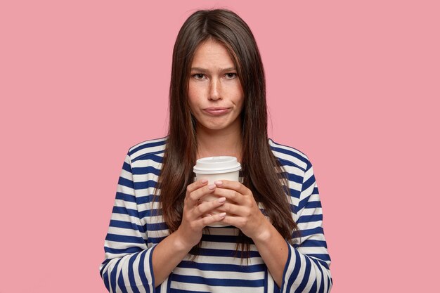 Красивая молодая женщина с грустным несчастным выражением лица держит одноразовый бумажный стаканчик, пьет кофе, чувствует себя расстроенной