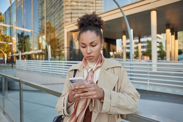아름다운 젊은 여성은 인터넷 연결이 좋은 응용 프로그램을 통해 온라인으로 휴대폰 확인 알림 채팅을 사용하여 진지한 표정을 지으며 도시 환경에 대한 비디오 포즈를 보고 있습니다.