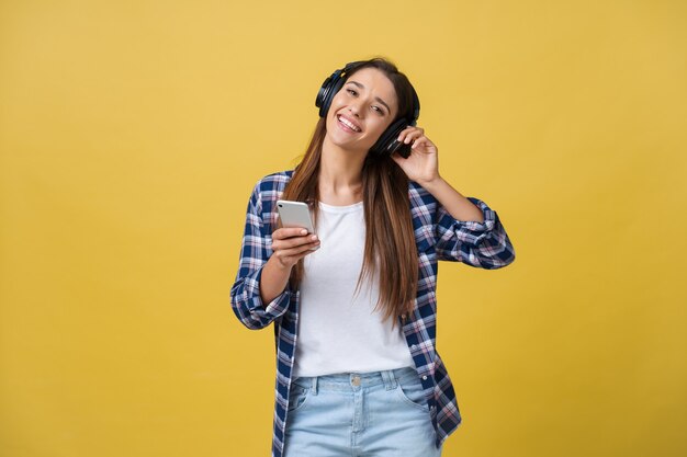 Beautiful young woman in headphones listening to music and dancing on yellow background.