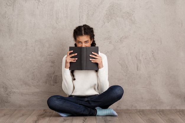 Free photo beautiful young woman hiding mouth behind book over beige wall