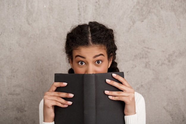 Beautiful young woman hiding mouth behind book over beige wall