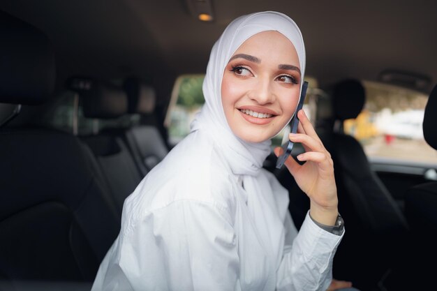 히잡을 쓴 아름다운 젊은 여성이 차에 앉아 전화 통화를 하고 있다