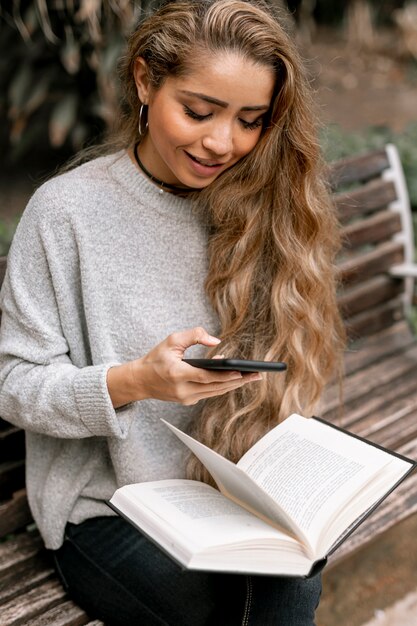 Beautiful young woman holding a book while checking her phone