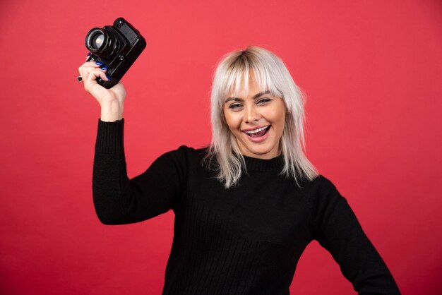 Beautiful young woman holding a camera while standing against red background. High quality photo