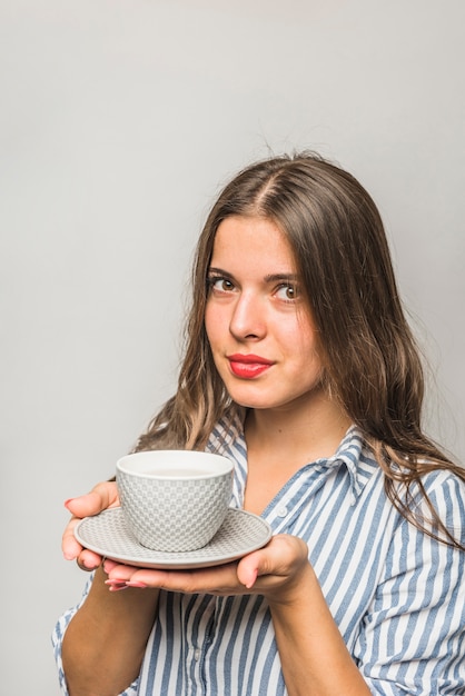 Foto gratuita bella giovane donna che tiene tazza grigia e piattino in mani contro il contesto grigio