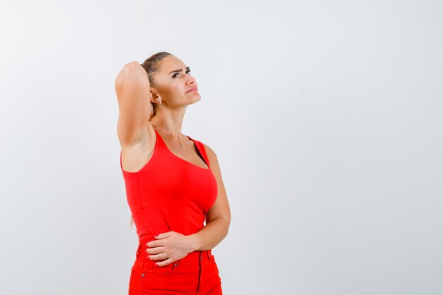 Beautiful young woman holding hand behind neck, looking up in red tank top, pants and looking pensive. front view.