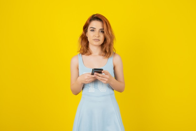 Beautiful young woman holding phone and looking at camera on yellow.