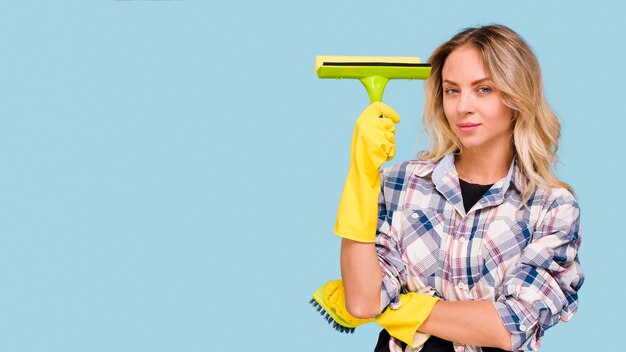 Beautiful young woman holding plastic wiper standing in front blue surface