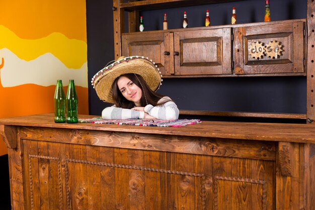 솜브레로를 입은 아름다운 젊은 여성이 멕시코 펍에서 맥주병을 들고 바 카운터에 기대었다
