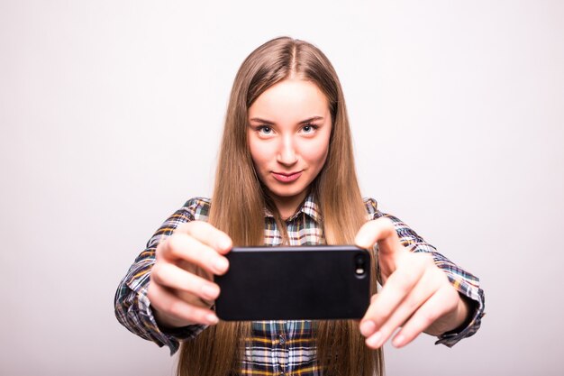 Красивая молодая женщина делает селфи фото со смартфоном, изолированным на белой стене