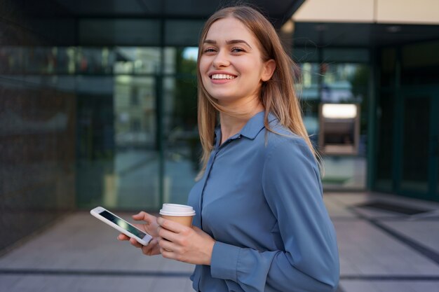 美しい若い女性は彼女のスマートフォンデバイスのアプリを使用してビジネスの建物の近くにテキストメッセージを送信しています