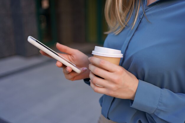 美しい若い女性は彼女のスマートフォンデバイスのアプリを使用してビジネスの建物の近くにテキストメッセージを送信しています