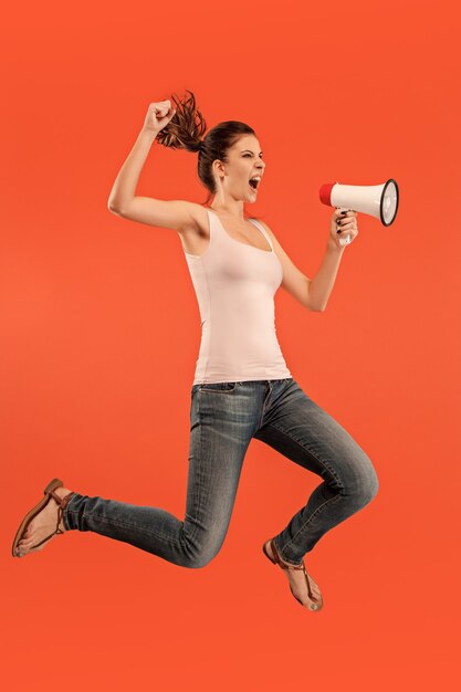 Beautiful young woman jumping with megaphone isolated over red background. Runnin girl in motion or movement. Human emotions and facial expressions concept