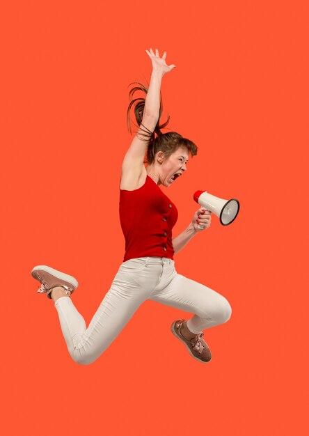 Beautiful young woman jumping with megaphone isolated over red background. Runnin girl in motion or movement. Human emotions and facial expressions concept