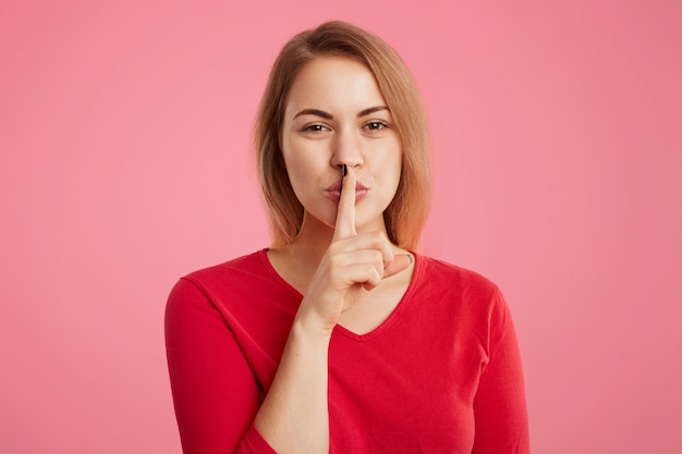 Beautiful young woman keeps fore finger on lips, shows silence sign, tries to keep personal information in secret