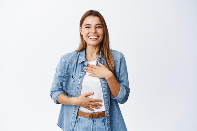 Beautiful young woman laughing and having fun, chuckle while holding hands on chest and smiling white teeth, express happiness and positive emotions, white