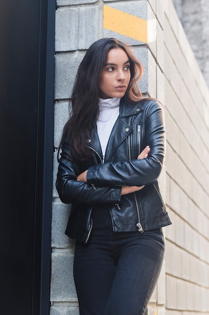 Beautiful young woman in leather jacket standing near the wall with her arms crossed