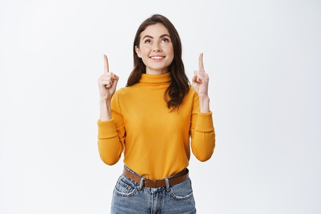 白い背景に黄色のセーターで立っている上に広告を表示して幸せな笑顔で見上げて上向きの美しい若い女性