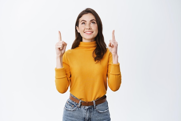 Beautiful young woman looking and pointing up with happy smile showing advertisement on top standing in yellow sweater against white background