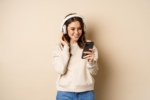 casuaに立っているヘッドフォンで音楽を聴いているスマートフォンでビデオを見ている美しい若い女性...