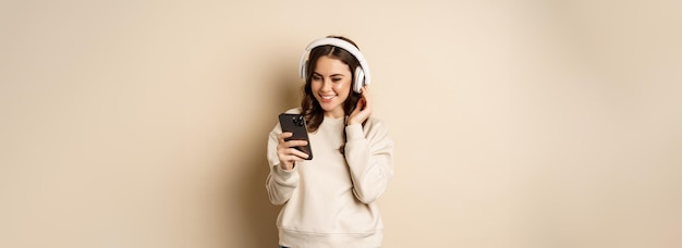 Free photo beautiful young woman looking at video on smartphone listening music in headphones standing in casual clothes over beige background