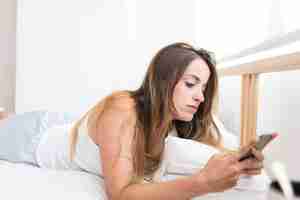 Free photo beautiful young woman lying on bed using smartphone