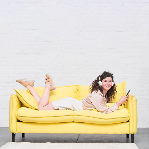 ヘッドホンで音楽を楽しむソファーに横たわっている美しい若い女性