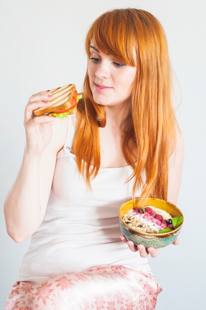 Beautiful young woman making choice between sandwich and oatmeal granola