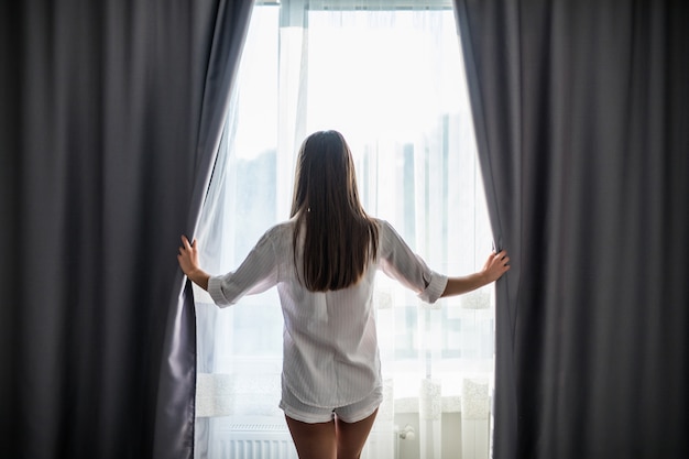 Beautiful young woman opening curtains and looking through the window
