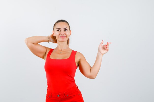 Beautiful young woman pointing at upper right corner, holding hand behind head in red tank top, pants and looking dreamy. front view.