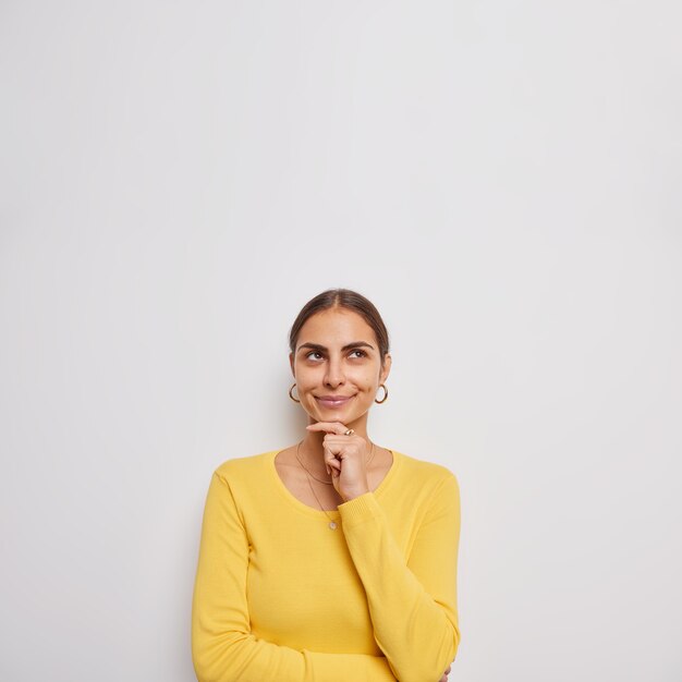 Beautiful young woman ponders something keeps hand on chin focused above being deep in thoughts wears casual yellow jumper isolated over grey wall