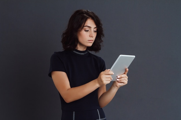 Beautiful young woman posing over grey wall and holding tablet in hands