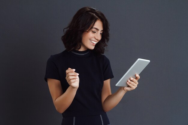 Beautiful young woman posing over grey wall and holding tablet in hands