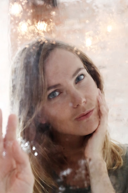 Beautiful young woman posing behind wet window