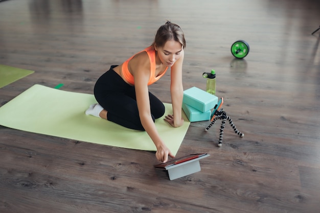 Beautiful young woman practicing yoga, is engaged with the teacher online via a tablet. Home sports concept.