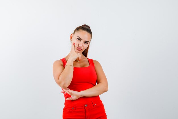 Beautiful young woman propping chin on hand in red tank top, pants and looking thoughtful. front view.