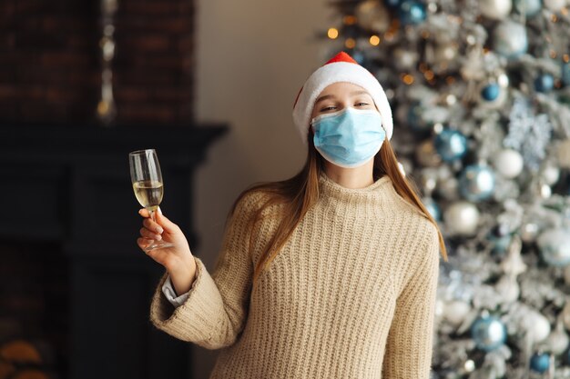 自宅でシャンパングラスと保護マスクの美しい若い女性。クリスマスのお祝い。コロナウイルスの制限の下でクリスマスを祝うという概念。