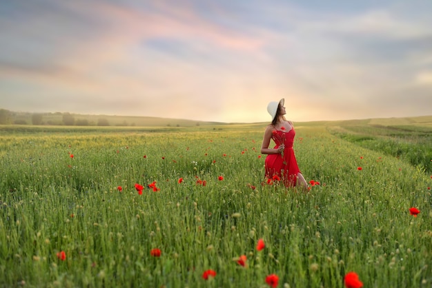 Beautiful young woman in red dress and white hat walks around field with poppies in a beautiful summ