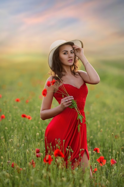 Beautiful young woman in red dress and white hat walks around field with poppies 