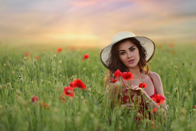 Beautiful young woman in red dress and white hat walks around field with poppies 