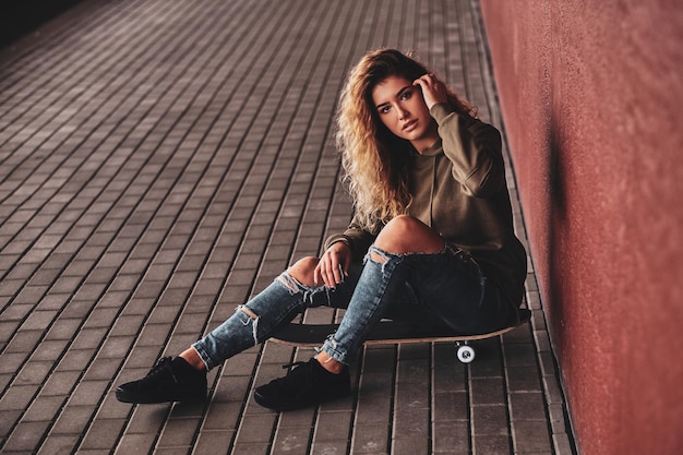 Bella giovane donna in jeans strappati è seduta sul suo skateboard in strada.