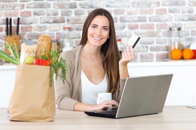 Beautiful young woman shopping with her card online in the kitchen.