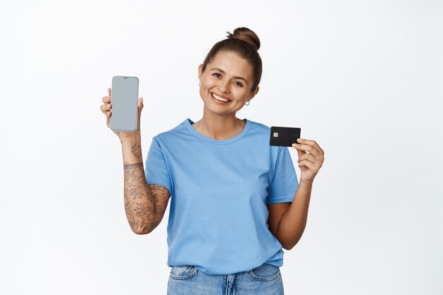 신용 카드, 앱 인터페이스가 있는 빈 전화 화면을 보여주는 아름다운 젊은 여성이 흰색으로 웃고 있습니다.