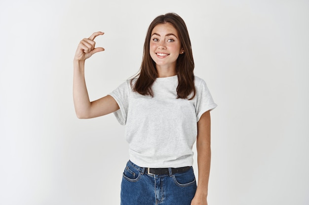 Bella giovane donna che mostra un piccolo, piccolo o piccolo gesto, con un sorriso felice, in piedi con una maglietta casual su un muro bianco