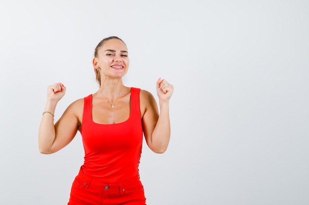 Beautiful young woman showing winner gesture in red tank top and looking happy , front view.