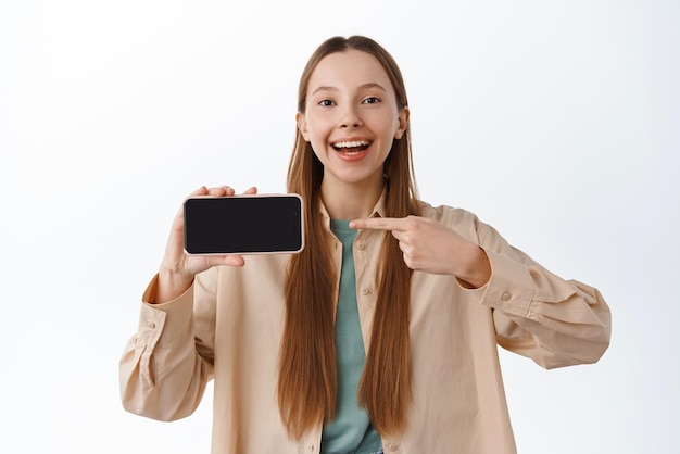 Beautiful young woman shows horizontal smartphone screen pointing at display and smiling recommending application standing over white background