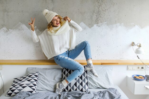 Beautiful young woman sitting in bedroom against wall wearing white sweater